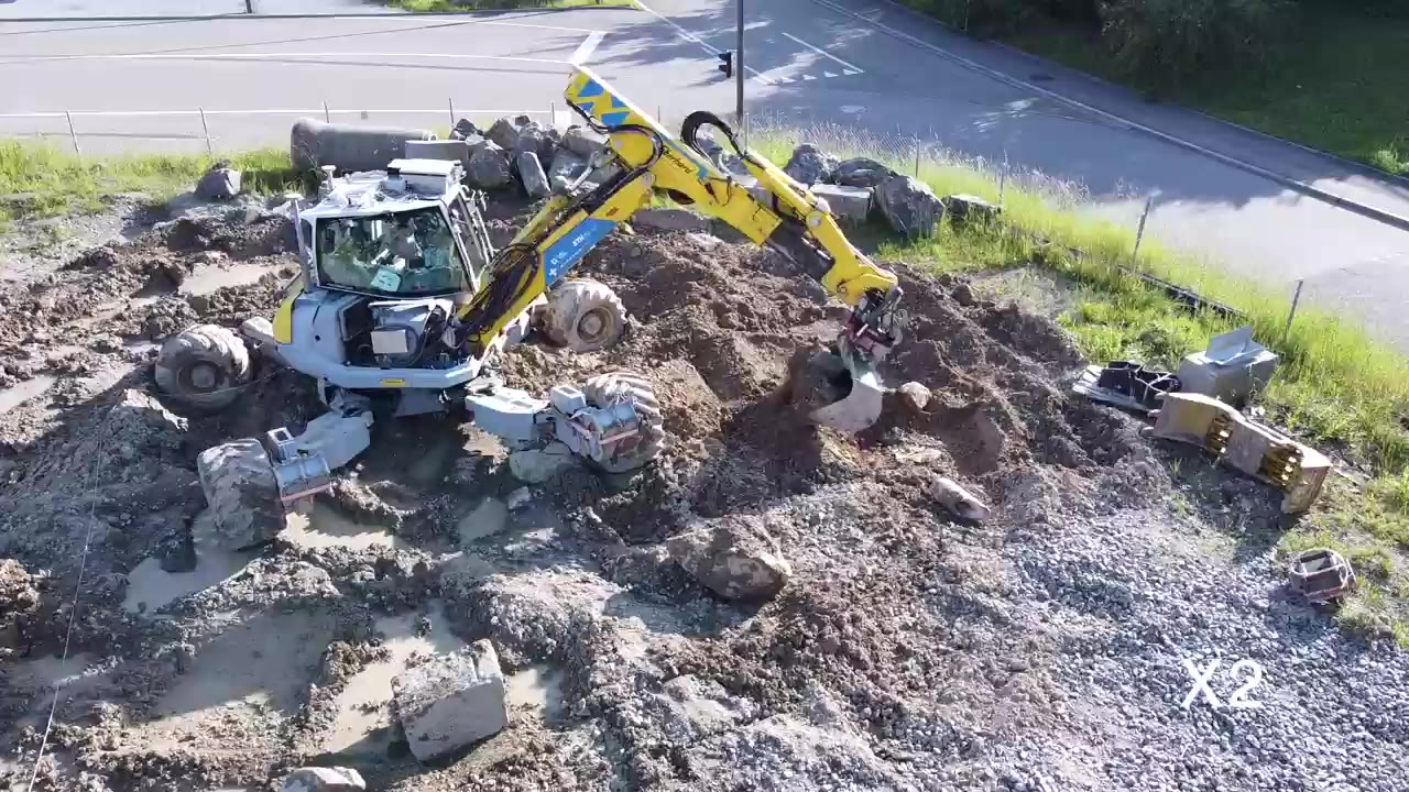 An overhead view of an excavator bucket contacting and lifting a boulder from rocky ground, shown at 2× speed in the source film.